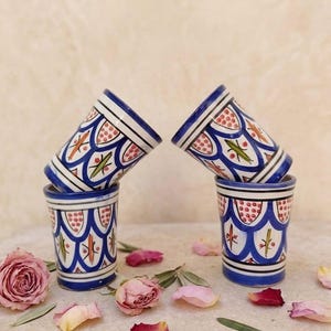 Peut inclure: Quatre tasses en céramique ornées de motifs peints à la main en bleu, rouge et vert. Deux tasses sont posées verticalement, tandis que deux autres sont équilibrées en angle au-dessus. Le design présente des lignes festonnées et florales.