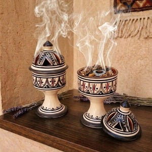 Peut inclure: Deux brûle-encens en céramique de couleur crème avec des bases sur pied et des couvercles amovibles. Les brûleurs présentent des motifs géométriques noirs, rouges et gris. L'un est fermé, l'autre ouvert, dégageant de la fumée blanche.