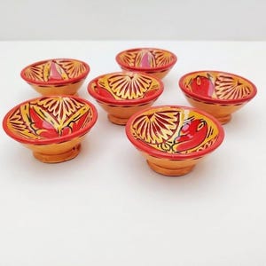 Peut inclure: Six petits bols en céramique peints à la main. Chaque bol présente une base arrondie et un motif vibrant avec des motifs rouges et dorés sur un fond orange clair. Les bols sont disposés sur une surface blanche.