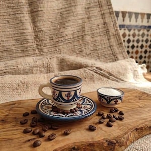 Peut inclure: Un ensemble tasse à espresso et soucoupe en céramique orné de motifs géométriques peints à la main en bleu et marron, accompagné d'un petit bol assorti rempli de sucre blanc, disposé sur une planche en bois parsemée de grains de café.