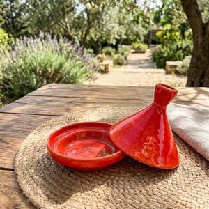 Peut inclure: Un tagine en céramique rouge vif, avec son couvercle conique légèrement entrouvert, repose sur un dessous de plat tissé. La finition brillante du tagine reflète la lumière du soleil. La base ouverte révèle l'intérieur.
