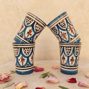 Peut inclure: Quatre tasses cylindriques en céramique ornées d'un motif peint à la main composé d'arches festonnées bleues, de points noirs et de lignes orange. Deux tasses sont posées verticalement, tandis que deux autres sont inclinées au-dessus.