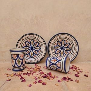 May include: Two ceramic cups and two matching saucers featuring a hand-painted blue, orange, and green floral pattern on a white background. The cups have a cylindrical shape, and the saucers have a wide rim with a repeating geometric border.