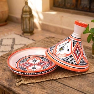 Può includere: Un tajine in ceramica con coperchio conico e base circolare abbinata. L'esterno presenta uno sfondo bianco con motivi geometrici dipinti a mano in arancione, nero e rosso, inclusi rombi, motivi floreali e bordi punteggiati.