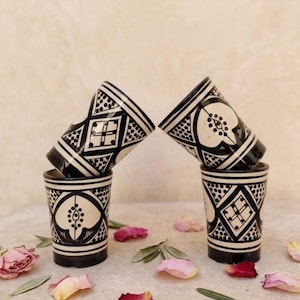 Może przedstawiać: Cztery ceramiczne kubki ułożone w dwie pary, każdy z kremową podstawą i skomplikowanymi, ręcznie malowanymi czarnymi geometrycznymi wzorami, w tym rombami, kropkami i motywami kwiatowymi, o gładkim, cylindrycznym kształcie.