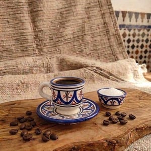 Peut inclure: Un ensemble tasse et soucoupe en céramique orné de motifs géométriques bleus et bruns, accompagné d'un petit bol assorti rempli de sucre blanc, le tout disposé sur une planche en bois avec des grains de café torréfiés éparpillés.