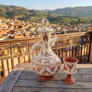 Può includere: Una brocca in ceramica con coperchio e un calice abbinato, entrambi con base color crema e motivi a foglia dipinti a mano in marrone rossastro. La brocca presenta un manico curvo, un collo stretto e un pomello appuntito sul coperchio, mentre il calice ha un piede.