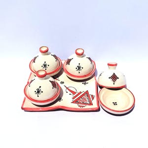 Puede incluir: Un juego de cuatro pequeños cuencos de cerámica blanca con bordes y tapas rojas, más una fuente. Cada cuenco tiene un diseño decorativo en negro y rojo. La fuente tiene un diseño a juego con formas geométricas rojas y negras.
