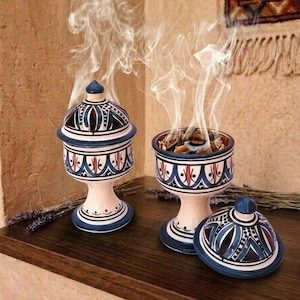May include: Two ceramic incense burners with cream, blue, and red geometric patterns. One burner is covered with a lid, while the other is open, emitting wisps of white smoke from burning incense. Both feature a pedestal base and a dome-shaped lid.