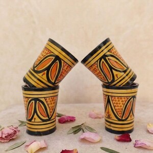 Peut inclure: Quatre tasses cylindriques en céramique disposées en deux piles de deux. Chaque tasse présente une base jaune avec des bandes horizontales noires et un motif répétitif de feuilles orange et noires. Le design inclut des points orange.