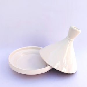 May include: A white ceramic tagine pot with a conical lid and a shallow, round base. The lid is slightly ajar, revealing the interior of the pot. The tagine is set against a plain white background.