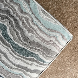 May include: Close-up of a decorative pillow with a wave-like pattern in shades of gray, white, and teal. The pillow has a textured appearance and a light-colored border. The pillow is placed on a brown surface.