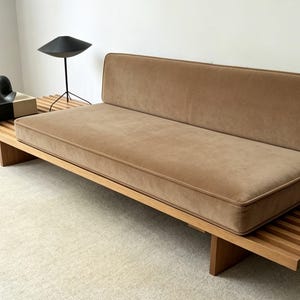 May include: A contemporary sofa featuring a light brown, velvet-style cushion and backrest. The sofa is set on a wooden slatted base with angled legs. A black lamp and decorative objects are on the side.