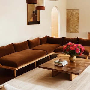 Op de afbeelding: Een moderne woonkamer met een grote, bruine hoekbank met houten frames. Een houten salontafel toont boeken en een vaas met roze bloemen. De kamer heeft een neutraal kleurenpalet met een crèmekleurig tapijt en kunstwerken.