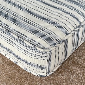 Puede incluir: Primer plano de un cojín cuadrado con un fondo color crema y rayas verticales azul marino. El cojín tiene una apariencia gruesa y acolchada con un borde a medida, lo que sugiere un artículo decorativo o funcional para la decoración del hogar.