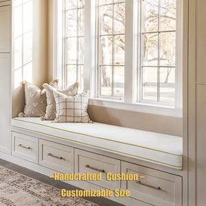 May include: A white window seat cushion with a gold trim, placed on a built-in bench with drawers. Decorative pillows with tassels and patterned designs are arranged on the cushion. The text on the image reads: "Handcrafted Cushion - Customizable Size."