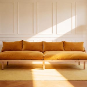 Op de afbeelding: Een mosterdgele bank met vier grote rugkussens, geplaatst op een licht houten frame en poten. Het staat op een gele vloerkleed. De achtergrond omvat een witte muur met rechthoekige panelen en een vloerlamp. De bank is ongeveer 200 cm lang.
