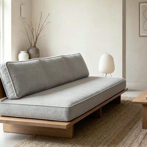 May include: A modern, low-profile daybed with a light gray cushion and a wooden frame. The cushion has a white trim. The daybed is placed on a wooden platform. The room has a neutral color palette.