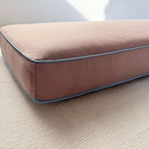 May include: A rectangular cushion with a muted rose-colored fabric and a light blue trim. The cushion is placed on a light-colored, textured surface. The edges are neatly sewn, suggesting a high-quality product.