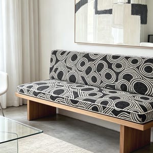 May include: A modern wooden bench with a black and white patterned cushion. The cushion features a repeating circular design. The bench has a light wood frame and is placed in a well-lit room with a painting on the wall.