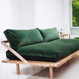 May include: A modern, green velvet futon sofa with a natural wood frame. The sofa features two large back cushions and a low profile design. The wooden frame has a minimalist aesthetic, with visible joinery and rounded legs. The overall style is contemporary and comfortable.