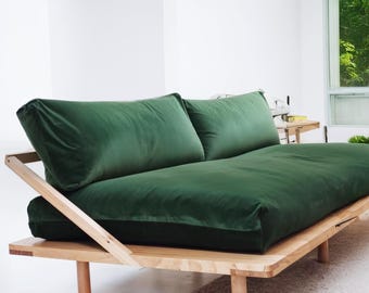 May include: A modern, green velvet futon sofa with a natural wood frame. The sofa features two large back cushions and a low profile design. The wooden frame has a minimalist aesthetic, with visible joinery and rounded legs. The overall style is contemporary and comfortable.