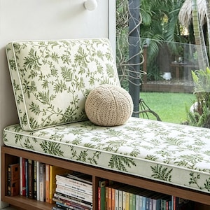 May include: A window seat with a cream-colored cushion and backrest, adorned with a green botanical pattern. A round, beige knitted pillow rests on the seat. The wooden base below houses a bookshelf filled with books.