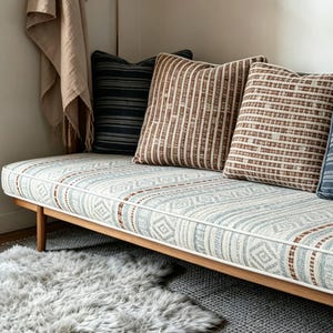 May include: A wooden daybed with patterned cushions and pillows in shades of brown, blue, and white. The pillows have striped and geometric designs. A fluffy gray rug sits on the floor in front of the daybed.