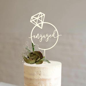May include: A small, round cake with a white frosting and a decorative topper. The topper is a ring design with the word "engaged" in script. The cake is decorated with a succulent and greenery, perfect for a wedding or engagement celebration.