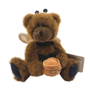 May include: A brown plush teddy bear with fuzzy fur, black paw pads, and two black antennae on its head. The bear has cream-colored wings on its back and holds a small tan plush honey pot with the text Bee and a bee graphic printed on it.