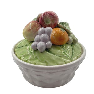 May include: A white ceramic bowl with a basket-weave texture on the exterior. The lid is shaped like a green leaf and is topped with a cluster of sculpted ceramic fruit, including a red apple, a yellow-green pear, a cluster of purple grapes, and an orange.