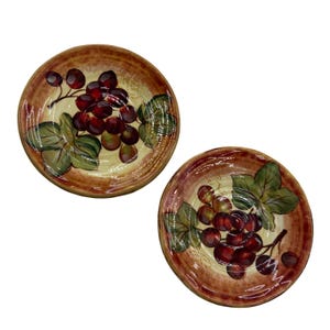May include: Two round ceramic plates featuring a hand-painted design of dark red grapes and green leaves. The plates have a cream-colored center and a wide, textured brown rim with a brushstroke finish, creating a rustic appearance.