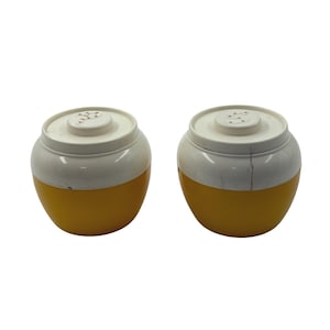 May include: Two round salt and pepper shakers with a two-tone design. The bottom half of each shaker is a solid yellow, while the top half and lid are white. The lids feature small perforated holes arranged in a letter S or P pattern.