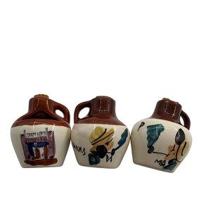 May include: Three ceramic jugs with brown glazed tops and cream-colored bases. The first jug features a painted building labeled CRAZY HORSE. The second jug shows a figure labeled PARKS. The third jug depicts a figure labeled MA, all with cork stoppers.