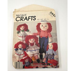 May include: A McCall's Crafts sewing pattern envelope, number 2447, featuring illustrations of Raggedy Ann and Andy dolls with red yarn hair, embroidered facial features, and various outfits including dresses, shirts, and pants with striped socks.