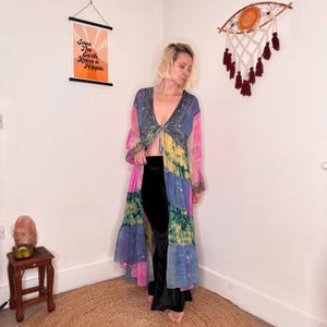 May include: A long, open-front duster cardigan featuring a patchwork design of tie-dye panels in shades of pink, yellow, blue, and green. The garment has long sleeves, a deep V-neckline with intricate silver embroidery, and a tie closure at the waist.