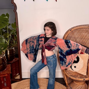 May include: A tie-front crop top featuring wide bell sleeves with a tie-dye pattern in shades of coral pink and dark navy blue, paired with blue denim jeans and a black tank top underneath, showcasing a flared silhouette and long flowing sleeves.
