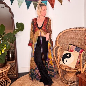 May include: A long, sheer, tie-front duster kimono featuring a vibrant, multi-coloured tie-dye pattern in shades of orange, yellow, and purple, worn over a black lace camisole and black, vertically-ribbed, wide-leg trousers.