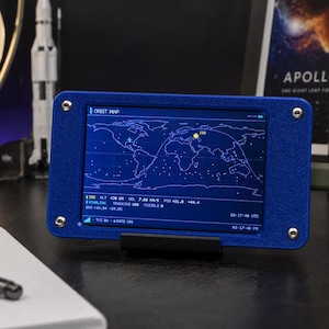 May include: A blue rectangular electronic device with a screen displaying an orbit map, world geography, and satellite tracking data including altitude, velocity, and coordinates. The device is secured with four silver screws at the corners.