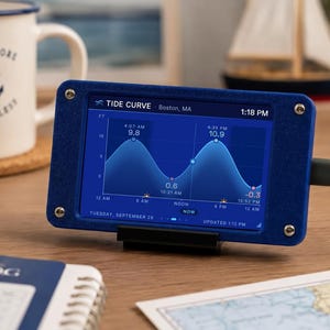 May include: A digital tide curve display device in a blue rectangular frame with four corner screws. The screen shows a graphical tide chart for Boston, MA, with time, date, and water levels in feet. A power cable is connected to the right side.