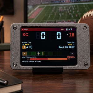 May include: A rectangular digital sports scoreboard device with a textured gray metal frame and four corner screws. The screen displays a live American football game between Kansas City and Tampa Bay, showing the score, down, and win probability.