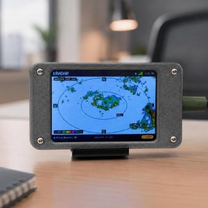 May include: A rectangular gray electronic device with a screen displaying a weather radar map. The screen shows a compass, precipitation intensity color scale, and the text RADAR, Olive Branch, MS, and Updated Just now. Four silver screws secure the frame.