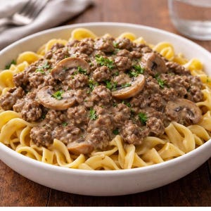 Könnte beinhalten: Eine weiße Schüssel gefüllt mit Eiernudeln, bedeckt mit einer cremigen braunen Soße, Hackfleisch, geschnittenen Pilzen und frischer Petersilie. Das Gericht steht auf einer Holzoberfläche, mit einer Gabel und einer Serviette im Hintergrund.