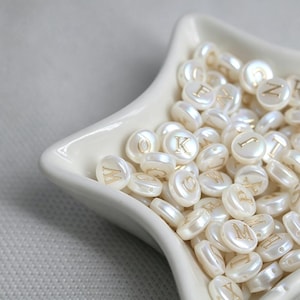 Pode incluir: Uma coleção de contas redondas, planas e esbranquiçadas com letras douradas, expostas numa tigela branca em forma de estrela. As contas são peroladas e têm um ligeiro brilho. As letras são em fonte serif.