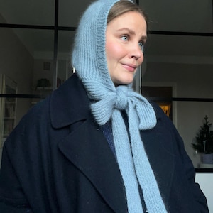 May include: A light blue knitted hood with long ties, worn over a dark navy coat. The hood is made of a soft, textured yarn and is tied in a bow at the neck. The overall look is cozy and stylish, perfect for cold weather.