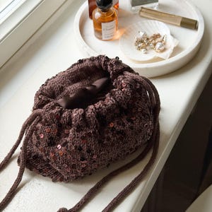 May include: A brown sequined drawstring bag with a knitted texture, on a white surface. Beside it is a white marble tray with perfume bottles, a seashell with jewellery, and a makeup tube.
