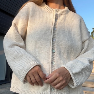Puede incluir: Cárdigan color crema con cuello redondo y mangas largas. El suéter de punto presenta botones azul claro y un diseño texturizado. Los puños y el dobladillo tienen un detalle acanalado. La prenda está hecha de un material suave y cálido.