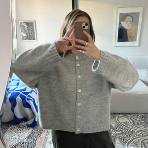 May include: A light grey button-down cardigan with a crew neck and long sleeves. The chunky knit sweater has white buttons. The person in the image is wearing dark trousers. The cardigan is a casual style.