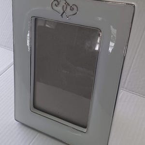 May include: A rectangular, white picture frame with a silver trim and a decorative silver heart design at the top. The frame has a glossy finish and a neutral color scheme, suitable for various decor styles.