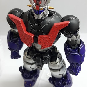May include: A robotic action figure featuring a black torso with a red hexagonal chest plate, silver limbs, and purple forearm and leg armor. The head has a silver crest, yellow eyes, and a blue face mask, standing on a clear plastic base.